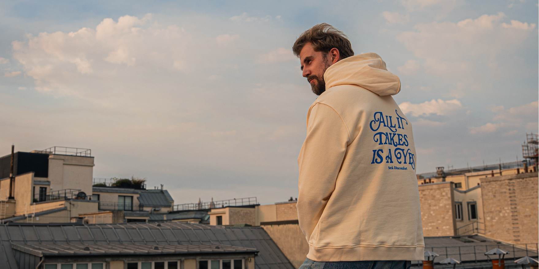 Thomas Brag wearing a cream-colored hoodie with blue text on the back stands on a rooftop overlooking city buildings under a cloudy sky.